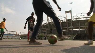 Teenagers play football outside an unfinished stadium