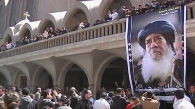 Mourners gathered in front of large banner showing image of Pope Shenouda III