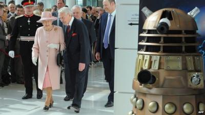 The Queen walks through MediaCity reception towards a Dalek