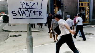 A man crossing the street in Sarajevo next to a sign warning of snipers