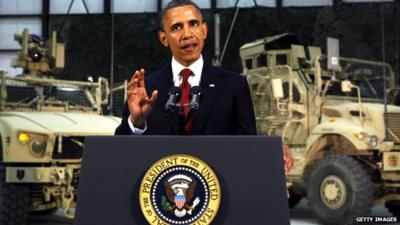 President Obama standing to give a speech with military trucks behind him