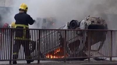 Firemen next to a burning upturned car