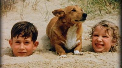 Prince Charles and Princess Anne as children buried in sand on beach with dog