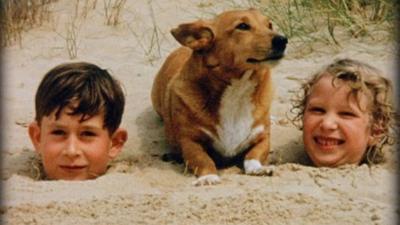 Prince Charles and Princess Anne as children buried in sand on beach with dog