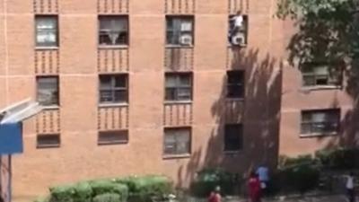 A young girl stands on an air conditioning unit outside of an apartment building window.