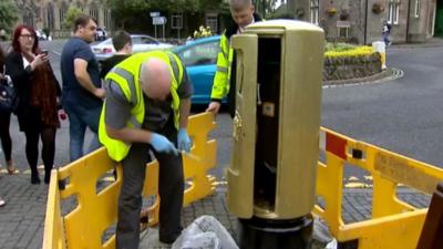 Crowd watches as a man paints a Royal Mail post box gold