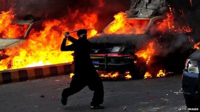 A Pakistani demonstrator brandishes a stick near burning police vehicles