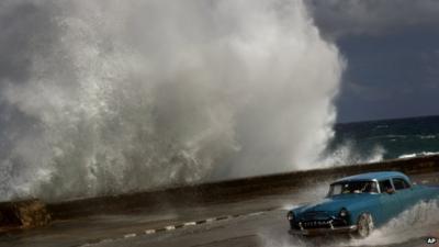 A driver maneuvers along a wet road as a wave crashes against the Malecon in Havana