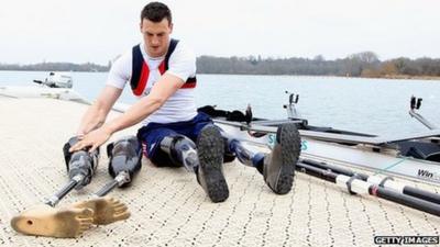 Paralympic rower Nick Beighton sits by the river next to his boat wearing prosthetic limbs
