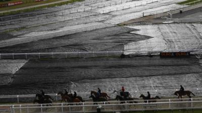 Horses and jockeys make their way past ground sheeting covering the course