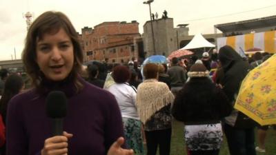 Julia Carneiro speaks to camera as Pope Francis gives a blessing in the background