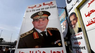 Egyptians walk behind a banner depicting U.S President Barak Obama, right, and a banner supporting Egyptian Army Chief Lt. Gen. Abdel-Fattah el-Sissi, center, with Arabic that reads, "God, keep this truthful honest man," in Cairo, Egypt