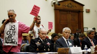 Protesters stand behind (from left) Chairman of the Joint Chiefs of Staff General Martin Dempsey, US Defence Secretary Chuck Hagel, and US Secretary of State John Kerry