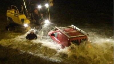 Waves crash around car, JCB and man