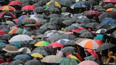 People take cover from the rain under umbrellas as they stand for the official memorial service for former South African President Nelson Mandela at the FNB Stadium