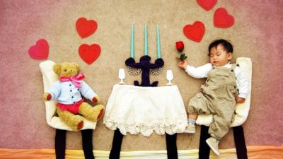 Photograph of staged scene of sleeping baby dining with a bear