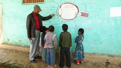 Man points to diagram of eye drawn on wall, children stand and watch