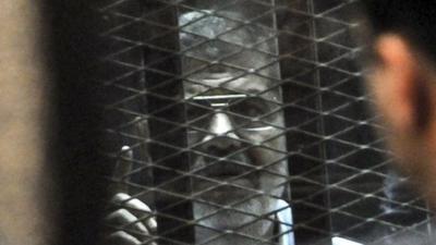 Mohammed Morsi speaks from a glassed-in defendant's cage during his trial over charges related to prison breaks