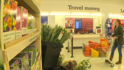 A lady pushes a trolley past a travel money counter in a supermarket