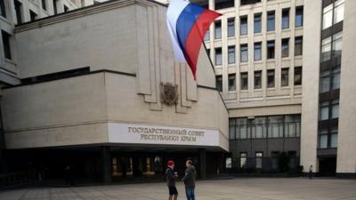 People stand under a Russian flag in front of the Crimean parliament bearing a new sign reading "State council of Crimean Republic" in central Simferopol