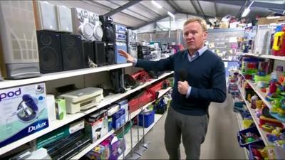 John Maguire in recycle shop next to shelves of electrical goods and toys