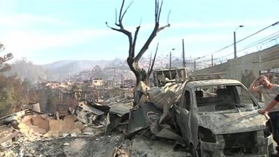 A man stands next to a car destroyed by fire