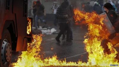 Protester in Guy Fawkes mask shielding self from flames near water cannon