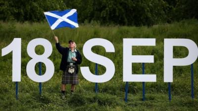 A man holding a Saltire flag stands next to giant letters which says "18 Sep".