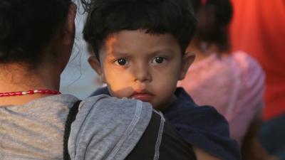 A Central American boy at the US-Mexico border is held in the arms of a woman.