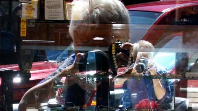 Patricia Lay-Dorsey photographing herself in the window of a shop, reflected in a mirror inside the shop