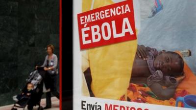 A woman walks an infant on a pushchair next to a bus stop banner reading "Emergency Ebola. Send doctors" in Spanish