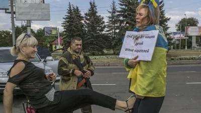 Irina, tied to a lamppost and draped in a Ukrainian flag, and a sign that reads (in Russian) "Ukrainian agent and baby-killer" being kicked by a woman