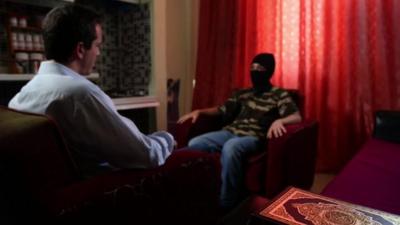 A 13-year-old boy who wishes to be named "Abu Hattab" sits wearing a black balaclava