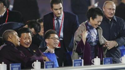 Vladimir Putin putting a coat around Peng Liyuan while seated alongside Xi Jinping and Barack Obama