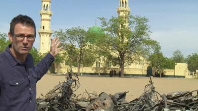 Will Ross pointing at mosque with pile of twisted metal in front of it