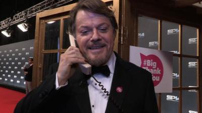 Eddie Izzard standing outside a phone box on the telephone