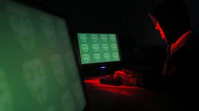 Hooded figure typing at a screen in a darkened room