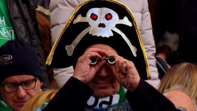 A fan in a pirate hat watches the rugby through binoculars