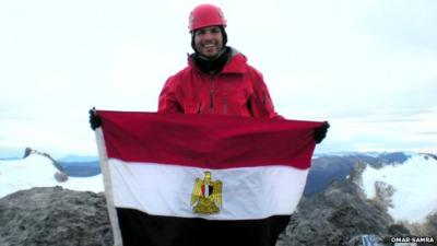 Omar Samra at the pinnacle of a mountain with a flag