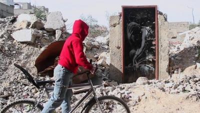 Boy on bike looks at door bearing Banksy artwork
