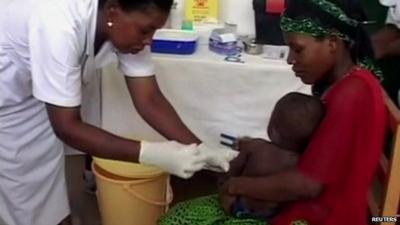 Nurse in Tanzania preparing to vaccinate a child (sitting on the mother's lap) against Malaria