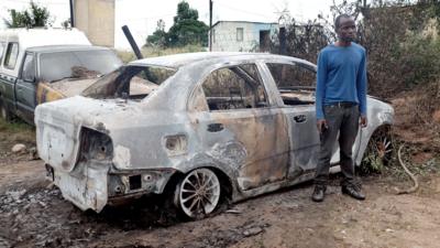 (File photo April 2015) A foreign national stands from Zimbabwe stands next to a car which has been attacked by a petrol bomb