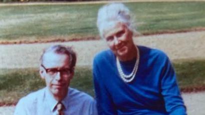 Man wearing glasses in pale blue shirt and tie sitting next woman with grey hair wearing blue dress