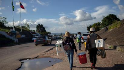 Pessoas carregam malas enquanto caminham em beira de estrada que liga Brasil e Venezuela