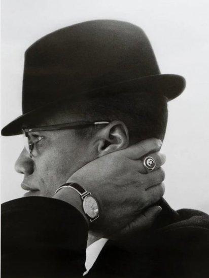 A black and white portrait of civil rights leader Malcolm X wearing a hat and displaying a star and crescent ring on his hand.