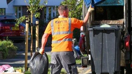A waste and refuse collector holding a bin bag and putting rubbish into a black wheelie bin.