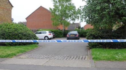 A police cordon in Frobisher Way, Shoeburyness. Police tape is blocking off the entrance to a small residential car park, which has three cars parked inside it. The area is surrounded by trees and bushes.