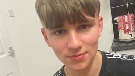 A boy wearing a black Nike-branded T-shirt faces the camera, there is a door and light switch visible behind him and a festive mug
