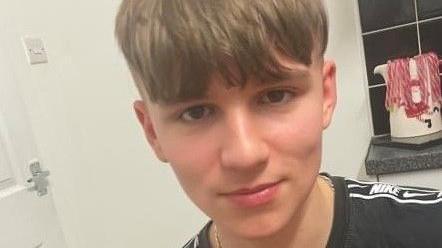 A boy wearing a black Nike-branded T-shirt faces the camera, there is a door and light switch visible behind him and a festive mug