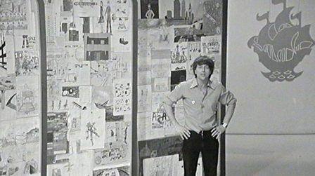 John Noakes, in the Blue Peter studio, standing in front of a board displaying several of the entries in the 'Ideas for the Year 2000' contest